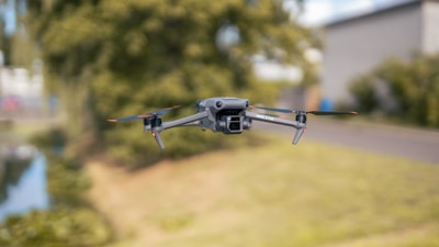 A drone hovers in mid-air against a blurred outdoor background with trees and a building. The drone is small, with a camera mounted on its front, and four propellers visible.