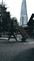 A person is pushing a hand truck loaded with boxes across a street in an urban setting. A tall, modern skyscraper is visible in the background, and trees line the sidewalk. The lighting suggests a cloudy day, casting shadows on the street.