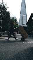 A person is pushing a hand truck loaded with boxes across a street in an urban setting. A tall, modern skyscraper is visible in the background, and trees line the sidewalk. The lighting suggests a cloudy day, casting shadows on the street.