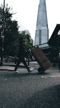 A person is pushing a hand truck loaded with boxes across a street in an urban setting. A tall, modern skyscraper is visible in the background, and trees line the sidewalk. The lighting suggests a cloudy day, casting shadows on the street.