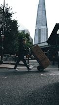 A person is pushing a hand truck loaded with boxes across a street in an urban setting. A tall, modern skyscraper is visible in the background, and trees line the sidewalk. The lighting suggests a cloudy day, casting shadows on the street.