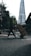 A person is pushing a hand truck loaded with boxes across a street in an urban setting. A tall, modern skyscraper is visible in the background, and trees line the sidewalk. The lighting suggests a cloudy day, casting shadows on the street.