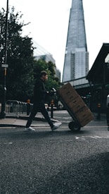 A person is pushing a hand truck loaded with boxes across a street in an urban setting. A tall, modern skyscraper is visible in the background, and trees line the sidewalk. The lighting suggests a cloudy day, casting shadows on the street.