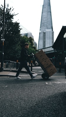 A person is pushing a hand truck loaded with boxes across a street in an urban setting. A tall, modern skyscraper is visible in the background, and trees line the sidewalk. The lighting suggests a cloudy day, casting shadows on the street.