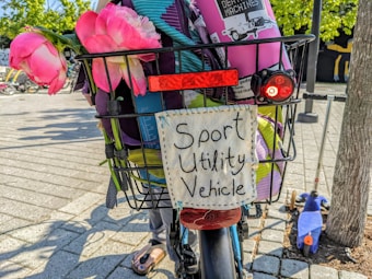 A bicycle basket filled with various items including pink flowers, cloth, and a sign that reads 'Sport Utility Vehicle'. The basket is attached to a bicycle with a purple background and a visible rear light. In the background, there are trees, a scooter, and a patterned pavement.