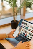 A person using a smartphone while sitting at a wooden desk with a laptop open displaying a grid of images. A potted plant is placed next to the laptop, adding a natural element to the workspace. The individual's hands are visible, and they are wearing a smartwatch.