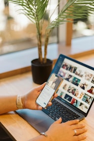 A person using a smartphone while sitting at a wooden desk with a laptop open displaying a grid of images. A potted plant is placed next to the laptop, adding a natural element to the workspace. The individual's hands are visible, and they are wearing a smartwatch.