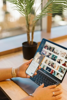 A person using a smartphone while sitting at a wooden desk with a laptop open displaying a grid of images. A potted plant is placed next to the laptop, adding a natural element to the workspace. The individual's hands are visible, and they are wearing a smartwatch.