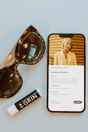 A mobile phone displaying a webpage about a summer cosmetics promotion sits on a light blue surface. Next to it, a large pair of brown sunglasses with a glossy frame and a black lip balm tube labeled 'GRIN' are placed. The screen shows a person wearing a yellow jacket against a wooden backdrop.