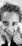 A close-up black and white photograph of a young child with curly hair smiling and looking directly into the camera. The child is standing on the grass, and the photographer's shoes are visible at the bottom.