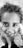 A close-up black and white photograph of a young child with curly hair smiling and looking directly into the camera. The child is standing on the grass, and the photographer's shoes are visible at the bottom.
