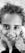 A close-up black and white photograph of a young child with curly hair smiling and looking directly into the camera. The child is standing on the grass, and the photographer's shoes are visible at the bottom.