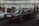 An indoor car exhibition with several vintage vehicles parked under industrial-style lighting. A dark-colored, classic sedan occupies the central position, with a visible license plate and polished surface reflecting the overhead lights. To the left, people are gathered around another car with its hood open. The background features a white brick wall with steel beams and stacked tires.