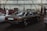 An indoor car exhibition with several vintage vehicles parked under industrial-style lighting. A dark-colored, classic sedan occupies the central position, with a visible license plate and polished surface reflecting the overhead lights. To the left, people are gathered around another car with its hood open. The background features a white brick wall with steel beams and stacked tires.