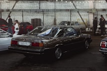 An indoor car exhibition with several vintage vehicles parked under industrial-style lighting. A dark-colored, classic sedan occupies the central position, with a visible license plate and polished surface reflecting the overhead lights. To the left, people are gathered around another car with its hood open. The background features a white brick wall with steel beams and stacked tires.