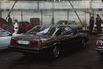 An indoor car exhibition with several vintage vehicles parked under industrial-style lighting. A dark-colored, classic sedan occupies the central position, with a visible license plate and polished surface reflecting the overhead lights. To the left, people are gathered around another car with its hood open. The background features a white brick wall with steel beams and stacked tires.