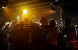 A group of young people stands in close proximity with their arms around each other, facing an audience under dim lighting. The crowd raises their arms and smartphones, capturing the moment. The atmosphere conveys a sense of unity and excitement, amplified by the warm tones of stage lights.