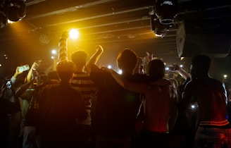 A group of young people stands in close proximity with their arms around each other, facing an audience under dim lighting. The crowd raises their arms and smartphones, capturing the moment. The atmosphere conveys a sense of unity and excitement, amplified by the warm tones of stage lights.