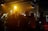 A group of young people stands in close proximity with their arms around each other, facing an audience under dim lighting. The crowd raises their arms and smartphones, capturing the moment. The atmosphere conveys a sense of unity and excitement, amplified by the warm tones of stage lights.