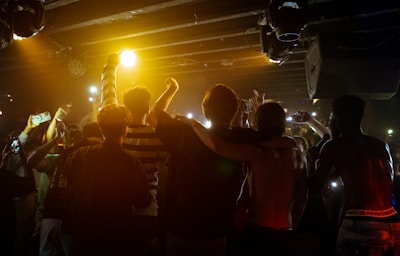 A group of young people stands in close proximity with their arms around each other, facing an audience under dim lighting. The crowd raises their arms and smartphones, capturing the moment. The atmosphere conveys a sense of unity and excitement, amplified by the warm tones of stage lights.