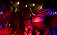 A vibrant concert scene with performers on stage, one of whom is singing into a microphone. The lighting is dramatic with bright stage lights overhead, casting red and yellow hues. The audience is energetic and engaged, with some people dancing and others capturing the moment on their phones.