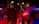 A vibrant concert scene with performers on stage, one of whom is singing into a microphone. The lighting is dramatic with bright stage lights overhead, casting red and yellow hues. The audience is energetic and engaged, with some people dancing and others capturing the moment on their phones.