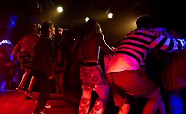 A vibrant concert scene with performers on stage, one of whom is singing into a microphone. The lighting is dramatic with bright stage lights overhead, casting red and yellow hues. The audience is energetic and engaged, with some people dancing and others capturing the moment on their phones.