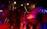 A vibrant concert scene with performers on stage, one of whom is singing into a microphone. The lighting is dramatic with bright stage lights overhead, casting red and yellow hues. The audience is energetic and engaged, with some people dancing and others capturing the moment on their phones.