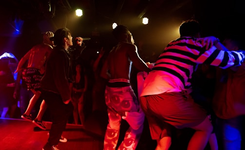 A vibrant concert scene with performers on stage, one of whom is singing into a microphone. The lighting is dramatic with bright stage lights overhead, casting red and yellow hues. The audience is energetic and engaged, with some people dancing and others capturing the moment on their phones.
