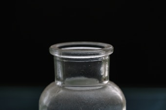 A close-up view of the top section of a clear glass bottle against a dark background. The bottle is cylindrical with a short neck and a slightly flared rim. The glass has a smooth texture and light reflections are visible on its surface.