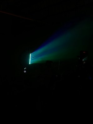 A dark setting with focused beams of colored light, primarily blue and green. The lights create a dramatic and atmospheric effect, possibly indicating a concert or club environment. A silhouette of a person holding a phone can be seen capturing the event.