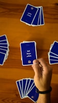 A hand holds a playing card with Arabic text printed on it. Several other cards are spread out on a wooden surface in different positions.