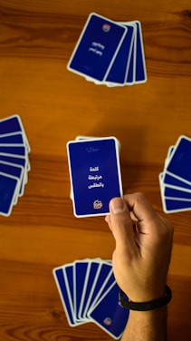 A hand holds a playing card with Arabic text printed on it. Several other cards are spread out on a wooden surface in different positions.