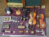 A collection of steampunk-themed objects laid out on a purple fabric surface. There are several watches with leather straps, vintage violin-like instruments with mechanical enhancements, and various metallic gadgets and jewelry pieces featuring gears. A notable black mask with metallic details, a skull inside a leather case, and an array of small accessories are also present. The items exhibit a blend of vintage artistry and mechanical complexity.