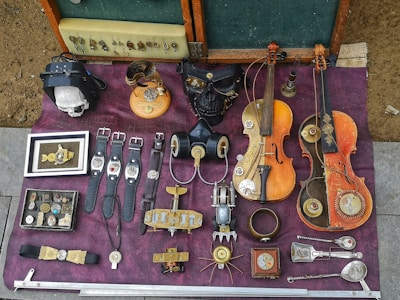 A collection of steampunk-themed objects laid out on a purple fabric surface. There are several watches with leather straps, vintage violin-like instruments with mechanical enhancements, and various metallic gadgets and jewelry pieces featuring gears. A notable black mask with metallic details, a skull inside a leather case, and an array of small accessories are also present. The items exhibit a blend of vintage artistry and mechanical complexity.