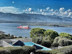 A scenic view of a body of water with a red ferry labeled 'Corsica Linea' sailing. In the foreground, there is a swimming pool with surrounding green foliage and a metal fence. The background shows a mountain range under a partly cloudy sky.