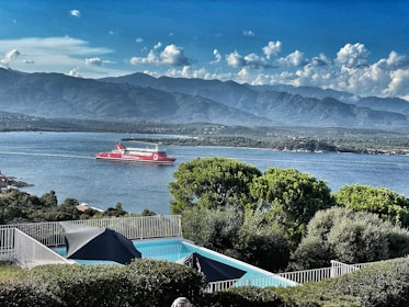 A scenic view of a body of water with a red ferry labeled 'Corsica Linea' sailing. In the foreground, there is a swimming pool with surrounding green foliage and a metal fence. The background shows a mountain range under a partly cloudy sky.