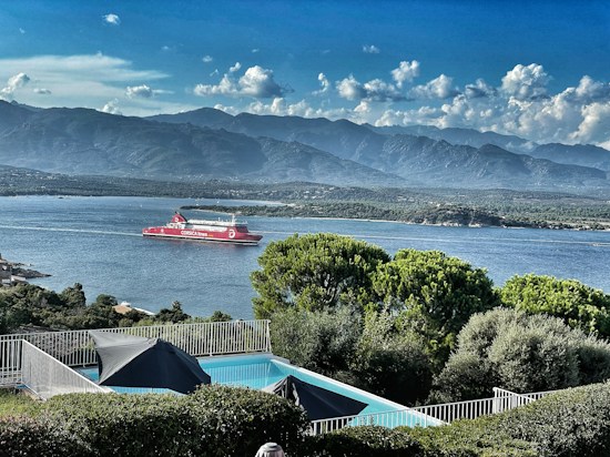 A scenic view of a body of water with a red ferry labeled 'Corsica Linea' sailing. In the foreground, there is a swimming pool with surrounding green foliage and a metal fence. The background shows a mountain range under a partly cloudy sky.