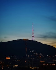 An illuminated radio or television tower stands atop a hill during evening or early night, surrounded by city lights at the base. The sky transitions from a deep blue at the top to a lighter horizon, indicating twilight.