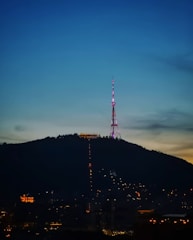 An illuminated radio or television tower stands atop a hill during evening or early night, surrounded by city lights at the base. The sky transitions from a deep blue at the top to a lighter horizon, indicating twilight.
