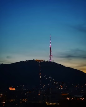 An illuminated radio or television tower stands atop a hill during evening or early night, surrounded by city lights at the base. The sky transitions from a deep blue at the top to a lighter horizon, indicating twilight.