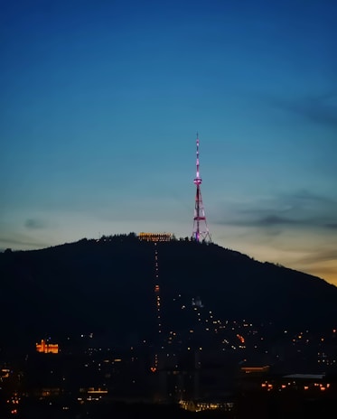 An illuminated radio or television tower stands atop a hill during evening or early night, surrounded by city lights at the base. The sky transitions from a deep blue at the top to a lighter horizon, indicating twilight.