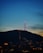 An illuminated radio or television tower stands atop a hill during evening or early night, surrounded by city lights at the base. The sky transitions from a deep blue at the top to a lighter horizon, indicating twilight.
