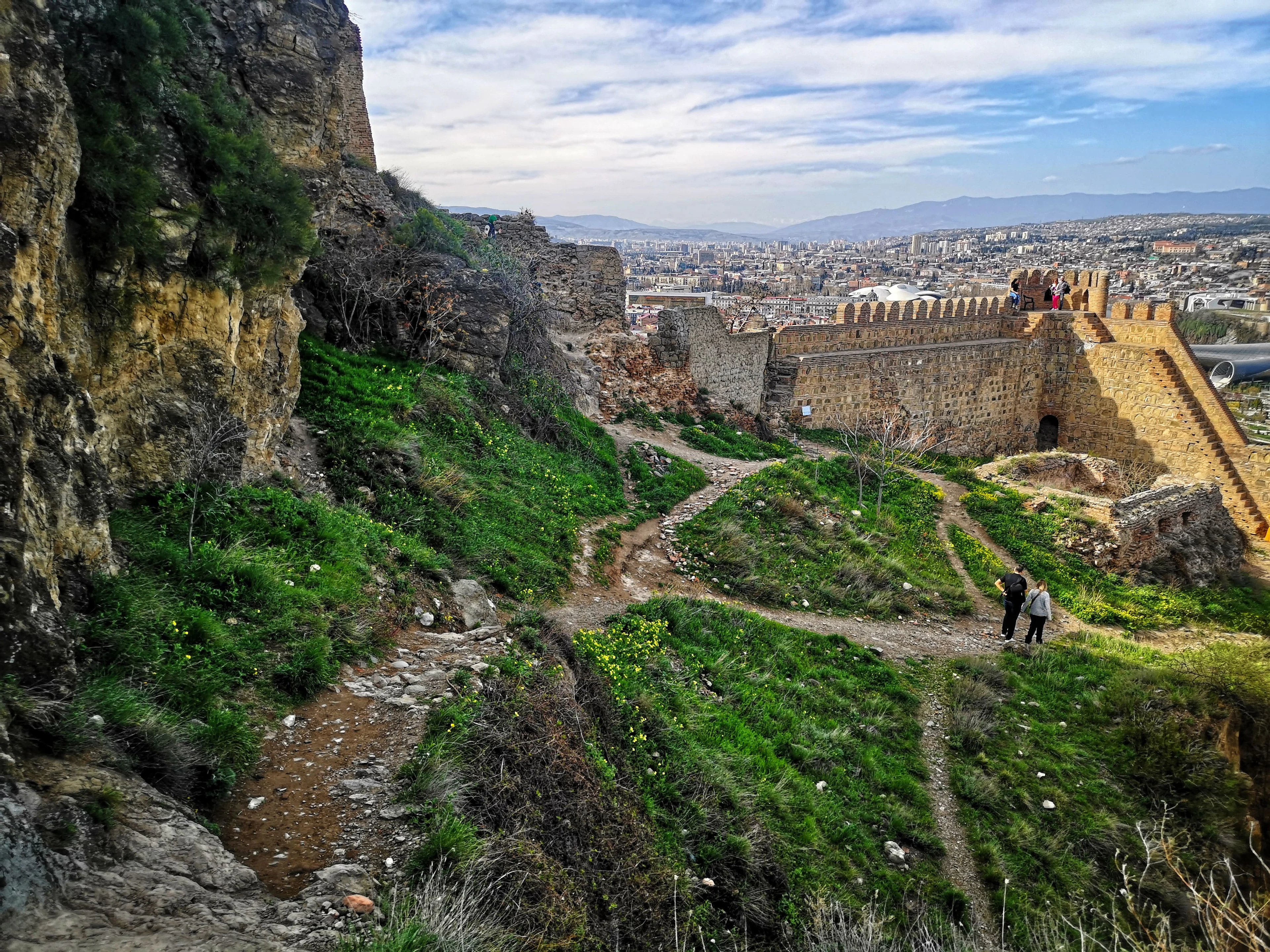 The ramparts of Narikala Fortress above Tbilisi in spring, crenellated brick walls and a round tower running along a ridge of bright green grass and yellow wildflowers, two walkers on the footpath and the city spreading to the mountains beyond