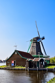 A traditional Dutch windmill is situated by a calm river, with a clear blue sky overhead. The windmill is accompanied by a rustic wooden building with a red and white door. The setting is peaceful and picturesque, showcasing a blend of natural and historical architecture.