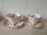 Two ornate teacups with matching saucers featuring a floral design with pink roses. Both cups and saucers have gold decorative accents and are placed on a light-colored countertop in front of a tiled backsplash.