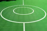 A vibrant green sports court featuring distinct white circular markings and straight lines. The court surface is composed of a textured grid pattern, and small leaves can be seen scattered on the surface.