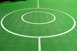 A vibrant green sports court featuring distinct white circular markings and straight lines. The court surface is composed of a textured grid pattern, and small leaves can be seen scattered on the surface.