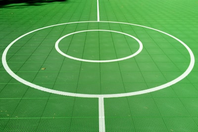A vibrant green sports court featuring distinct white circular markings and straight lines. The court surface is composed of a textured grid pattern, and small leaves can be seen scattered on the surface.