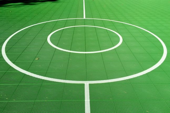 A vibrant green sports court featuring distinct white circular markings and straight lines. The court surface is composed of a textured grid pattern, and small leaves can be seen scattered on the surface.